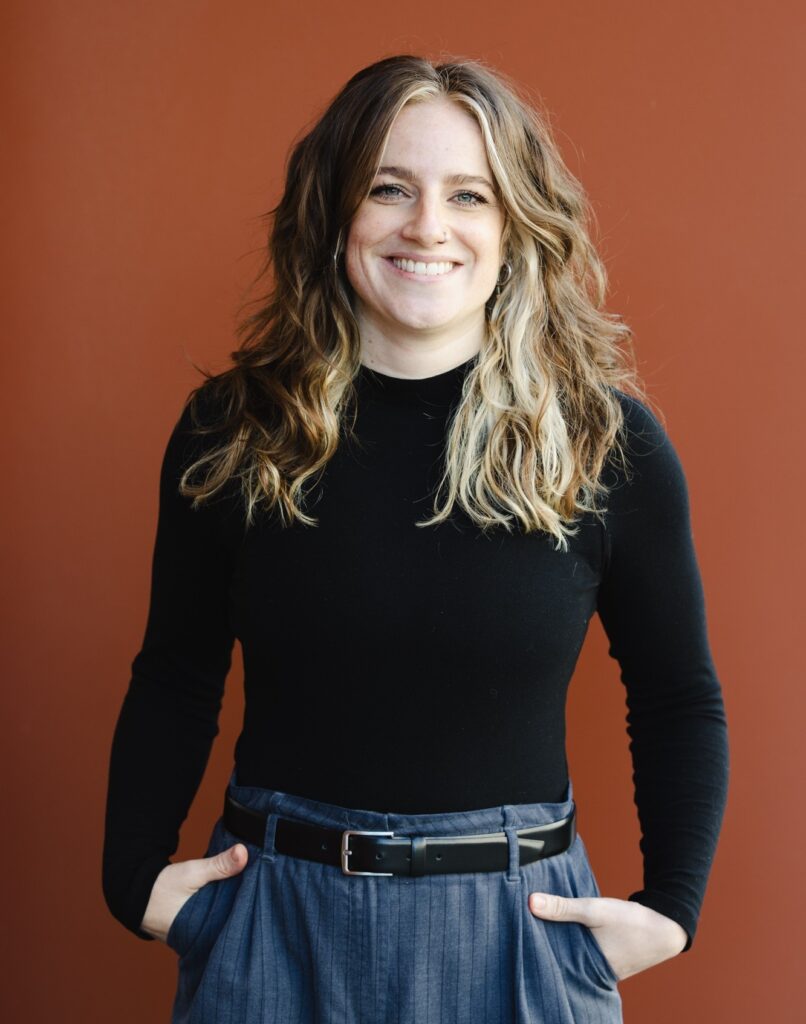 Rebekka hat lange braune Haare mit hellen Strähnen.
Sie lächelt mit offenem Mund in die Kamera.
Sie trägt ein langärmliches schwarzes Oberteil.